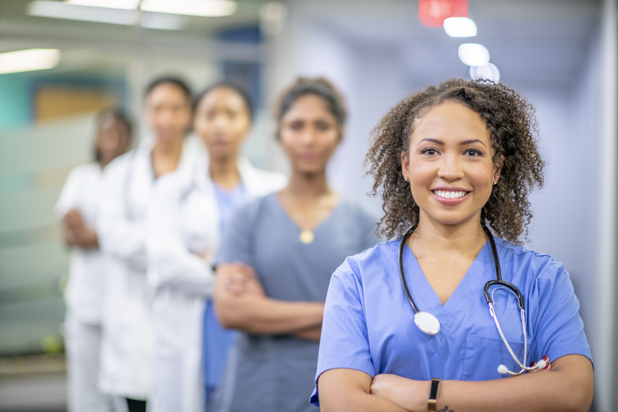 A beautiful female doctor of African descent is standing in front of her coworkers at the hospital. She is smiling widely at the camera.